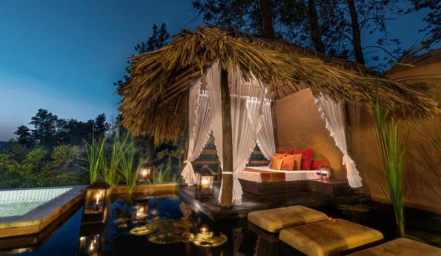 Luxurious outdoor cabana with white drapes under a thatched roof, surrounded by water and lanterns, creating a serene, romantic ambiance.

