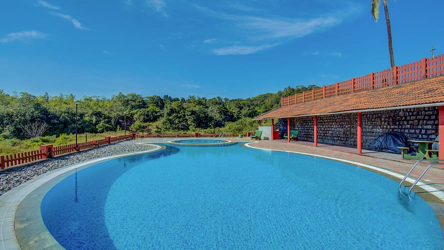 Large outdoor swimming pool with clear blue water, surrounded by greenery, a stone wall, and a shaded pavilion with red-tiled roofing.