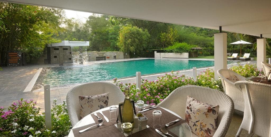 Poolside dining area with cozy wicker chairs, floral cushions, and a serene view of a large swimming pool surrounded by lush greenery.